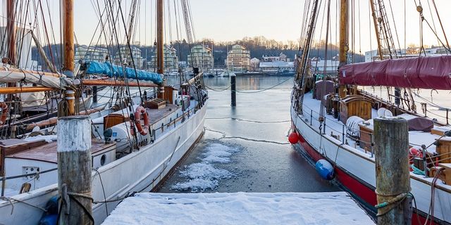 Wintercamping an der Ostsee: An einem verschneiten Dock in Flensburg liegen zwei Boote an. Das Wasser ist zum Teil zugefroren und gegenüber vom Hafen ist eine Häuserreihe sowie weitere Boote am Ufer.