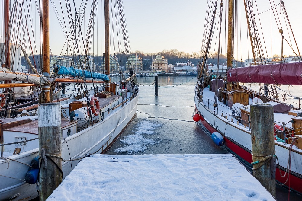 Wintercamping an der Ostsee: An einem verschneiten Dock in Flensburg liegen zwei Boote an. Das Wasser ist zum Teil zugefroren und gegenüber vom Hafen ist eine Häuserreihe sowie weitere Boote am Ufer.