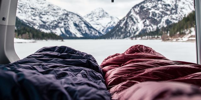Zwei Menschen liegen in Schlafsäcken, die aus der geöffneten Heckklappe eines Campers herausragen. Der Camper ist umgeben von einer verschneiten Berglandschaft.