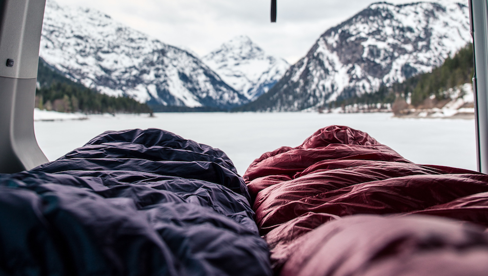 Zwei Menschen liegen in Schlafsäcken, die aus der geöffneten Heckklappe eines Campers herausragen. Der Camper ist umgeben von einer verschneiten Berglandschaft.