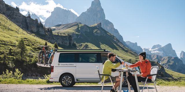 Ein Pärchen in Sportklamotten sitzt beim Camping Urlaub mit Fahrrad vor ihrem Camper in der Bergen und frühstückt vor einer Bergkulisse und grünen Wiesen