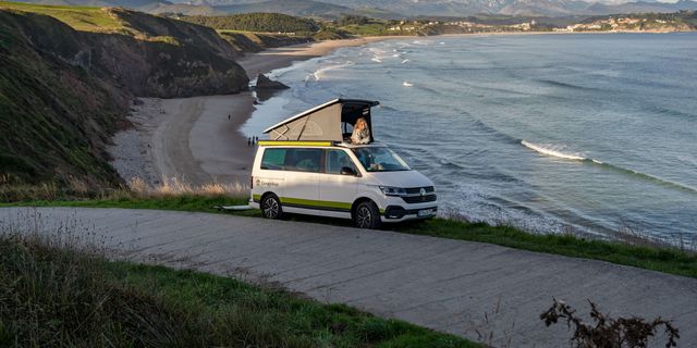 VW T6.1 California Beach Ocean steht vor Strand, mit Frau im Aufstelldach