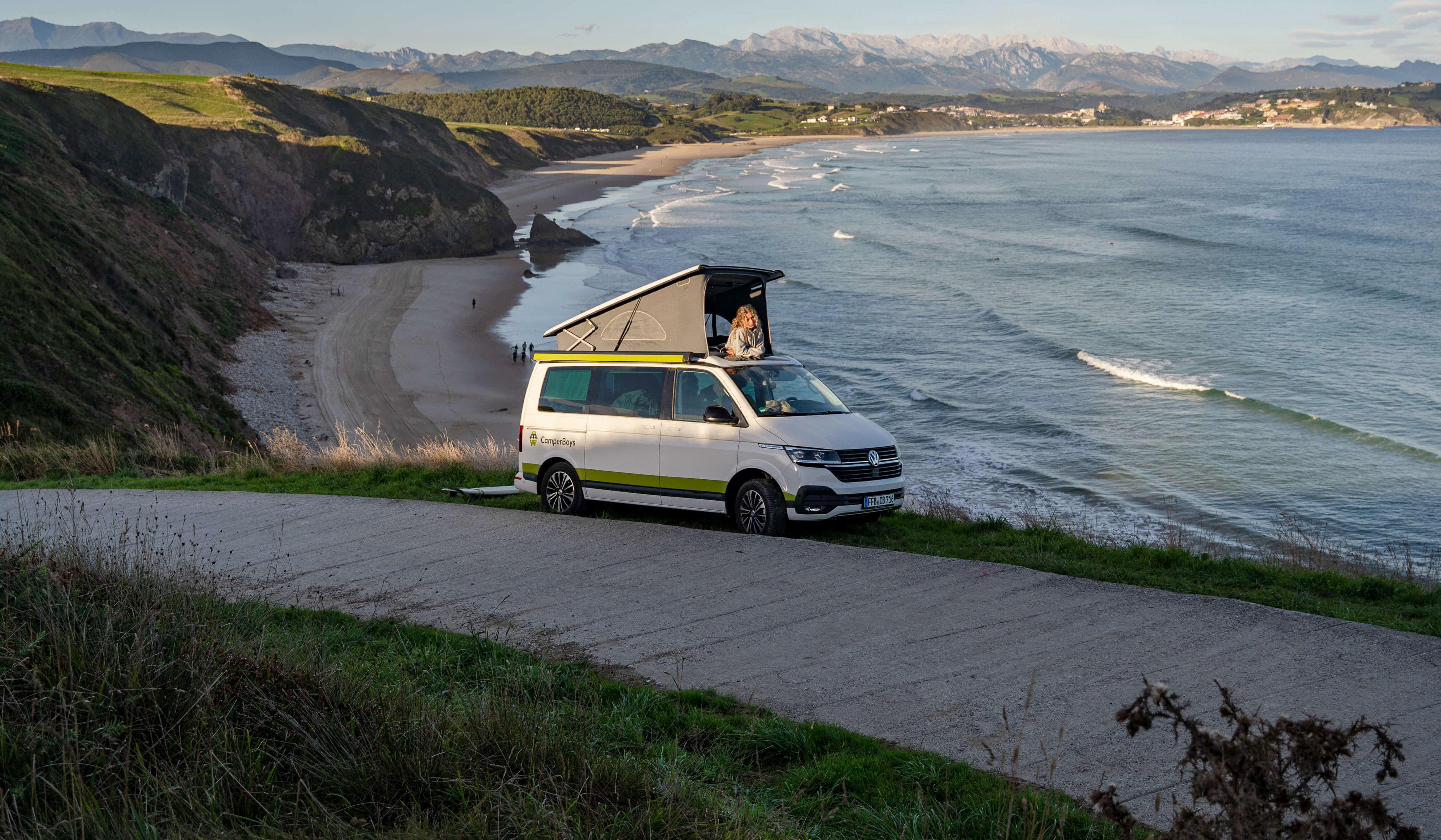 VW T6.1 California Beach Ocean steht vor Strand, mit Frau im Aufstelldach