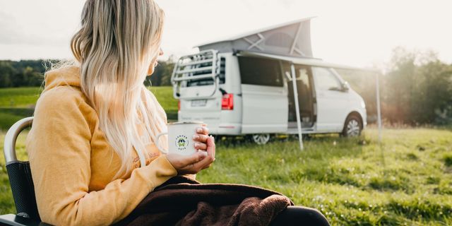 Blonde Frau sitzt auf einem Campingstuhl vor einem CamperBoys Camper und hat eine CamperBoys Tasse in der Hand.