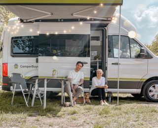 Ein Mann und eine Frau sitzen vor ihrem mit Lichterketten verzierten CamperBoys Camper im Grünen in Südholland und lachen in die Kamera 