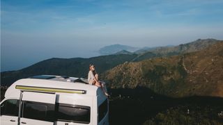 Eine Frau sitzt auf dem Dach eines VW Grand California, im Hintergrund sieht man Berge und Meer.
