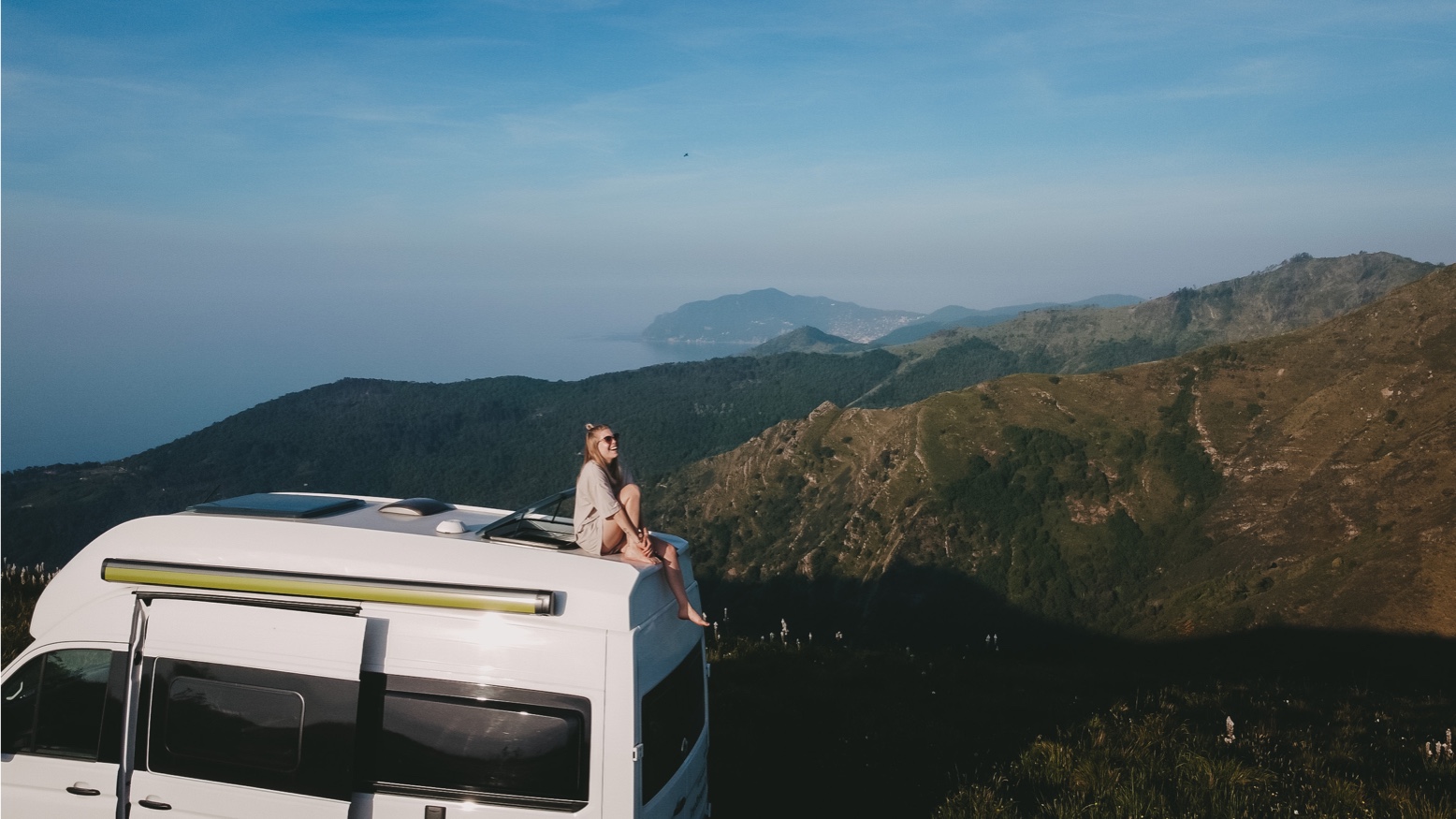 Frau auf Dacht von VW Grand California, im Hintergrund Berge und Meer