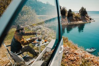 Mann frühstückt neben Camper an einem See
