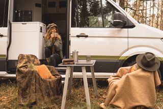 Frau beim Campen im Herbst sitzt auf der Treppe ihres Campers im Wald in herbstlichem Setting und trinkt einen Kaffee
