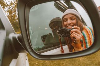 Eine Frau macht mit einer Kamera ein Foto von sich selbst im Rückspiegel eines Campers.