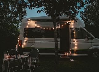 Ein CamperBoys VW Grand California steht auf Gras, davor ein gedeckter Campingtisch. Es ist dunkel und der Camper ist mit leuchtenden Lichterketten dekoriert.