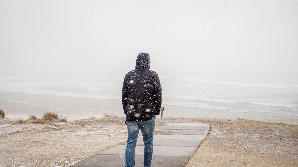 Ein Mann von hinten in dunkler Jacke. Er steht am Strand und es schneit.