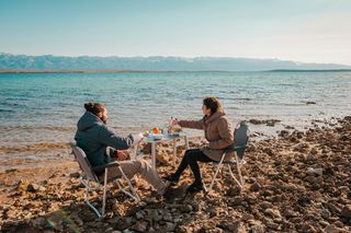 Mann und Frau beim Essen an steinigem Strand vor einer Kulisse aus Bergen und Meer 