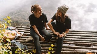 Zwei junge Frauen sitzen während ihres Riegsee Campings auf einem Steg, um beim wandern eine Pause zu machen und lachen sich an