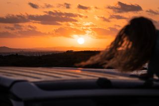 Die Sonne geht unter und eine Frau schwingt sich auf das Dach eines CamperBoys Vans, um den Sonnenuntergang zu beobachten.