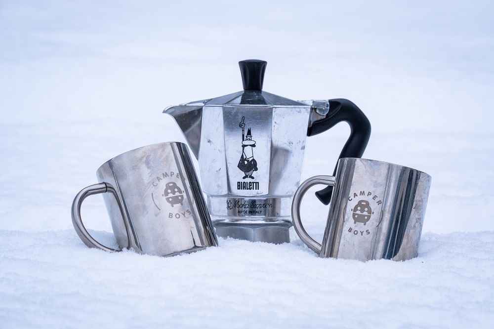 Eine Espressokanne und zwei silberne Becher mit CamperBoys Logo stehen im Schnee.
