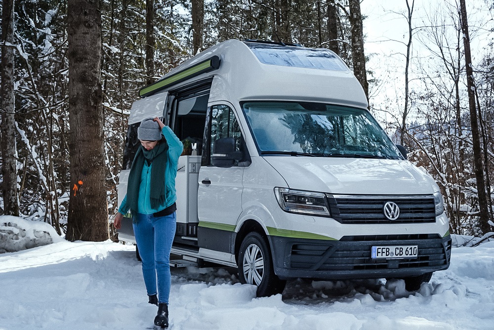 Ein VW Grand California steht im Schnee auf einem Waldgrundstück. Eine Frau in Winterkleidung geht neben ihm.